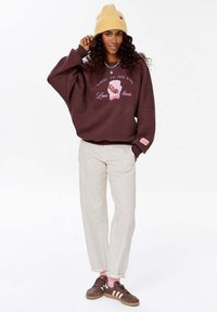 Žena v žlutém klobouku, hnědém svetru s grafikou psí židle, světle béžových kalhotách, růžových ponožkách a hnědých teniskách pózuje s jednou rukou u klobouku.