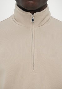 Beige zip-up-pullover i texturerat tyg med hög krage; har en silverfärgad dragkedja vid halsen och rena sömdetaljer.
