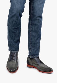 Scarpe da dress in pelle grigia con dettagli perforati, arricchite da una linguetta in tessuto blu, abbinate a un paio di jeans in denim scuro. Accenti rossi e blu sulla suola.