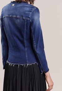 Jeansjacka i mörkblått med en figurnära design, fransad nederkant och råa kanter. Har kontrasterande sömmar och en metallmärke på baksidan.