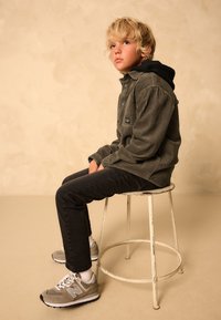 Veste-chemise en velours côtelé gris avec capuche noire, jean noir et baskets New Balance gris. Le modèle est assis sur un tabouret en métal blanc usé.