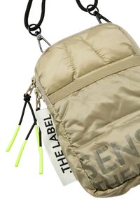 Gepolsterte, beige Umhängetasche mit glatter Oberfläche, verstellbarem schwarzen Tragegurt, neon grünen Reißverschlussanhängern und einem weißen Etikett mit schwarzem Text.