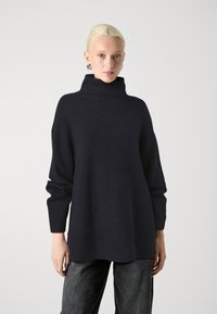 Temno modra oversized pulover z visokim ovratnikom, rebrastimi manšetami in robom, izdelan iz mehkega pletenega materiala. Opazna subtilna tekstura.