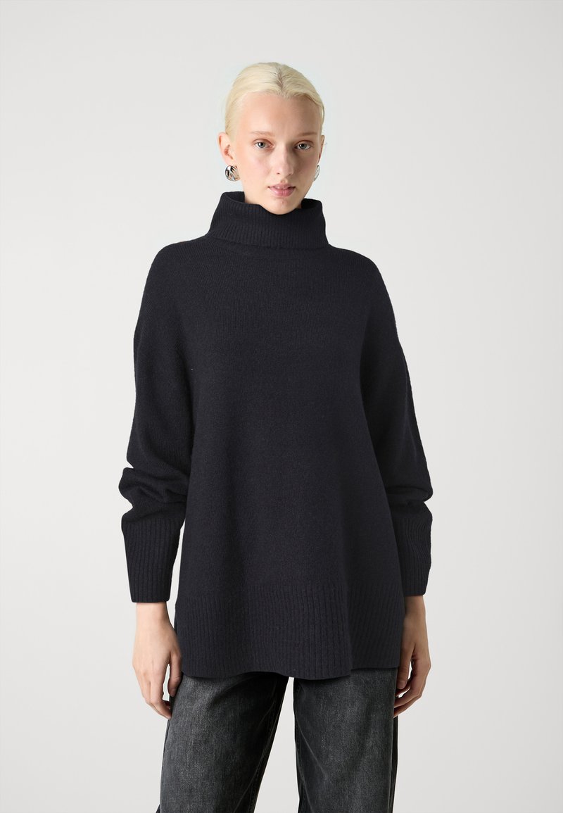 Temno modra oversized pulover z visokim ovratnikom, rebrastimi manšetami in robom, izdelan iz mehkega pletenega materiala. Opazna subtilna tekstura.