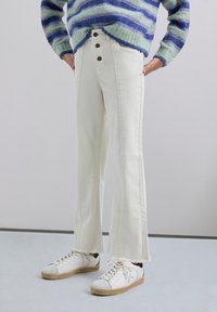 Jeans blancos acampanados con cuatro botones delanteros, bolsillos frontales y dobladillos deshilachados, combinados con zapatillas blancas. El suéter presenta rayas azules y verdes.