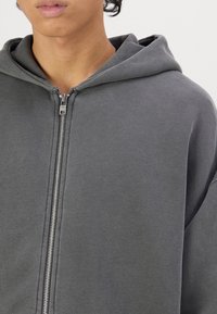 Grå zip-up hoodie med en rymlig huva, tillverkad av mjukt tyg; har en central dragkedja och något sänkta axlar. Inga mönster eller detaljer.