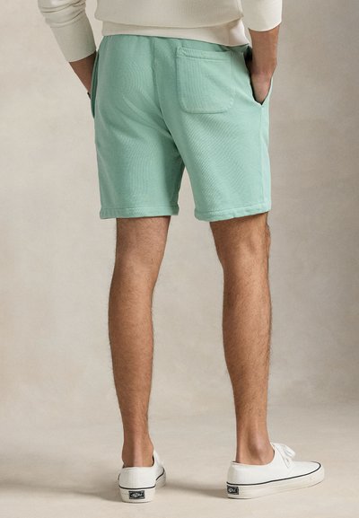 Πράσινα σορτς μέχρι το μέσο του μηρού, κατασκευασμένα από απαλό υλικό, με τσέπη στο πίσω μέρος και ρεβέρ στο τελείωμα. Φοριούνται με λευκά πάνινα παπούτσια.