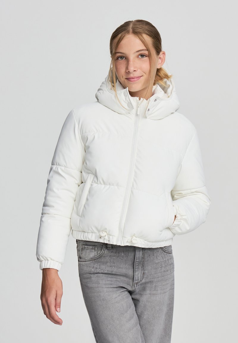 Biała kurtka puchowa z odpinanym kapturem, zamek błyskawiczny, elastyczne mankiety i dwie boczne kieszenie. Nosi się ją na szarych dżinsach.