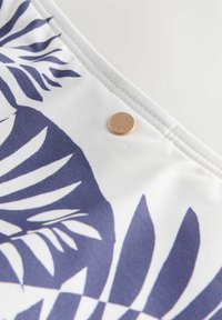 Fehér szövet kék trópusi levélmintával és egy kis, kerek arany fém címkével a varrott varrás közelében.