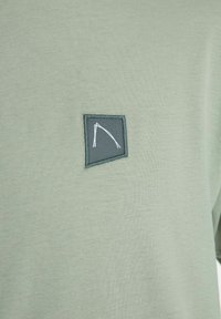 Tricou verde deschis dintr-un material texturat. Prezintă o emblema dreptunghiulară de culoare verde închis cu cusături albe, care afișează un design geometric minimalist.