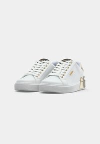 Une paire de baskets blanches basse avec des œillets en or, un logo doré sur les côtés et des accents de talon en or métallisé, présentée sur un fond blanc.