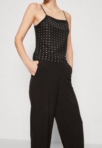 Zwarte satijnen camisole met dunne bandjes, versierd met zilveren studs in een patroon, gecombineerd met zwarte wijde broek, gepresenteerd in een staande pose.