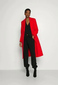 Vrouw staat gekleed in een helder rode lange jas over een zwarte V-hals top, zwarte broek en zwarte enkellaarsjes tegen een eenvoudige lichte achtergrond.