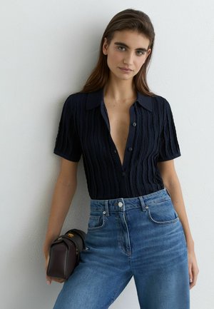 Kobieta w czarnej, teksturowanej koszuli z krótkim rękawem i guzikami oraz niebieskich, wysokich dżinsach, trzymająca ciemnobrązową torebkę na tle białej ściany.