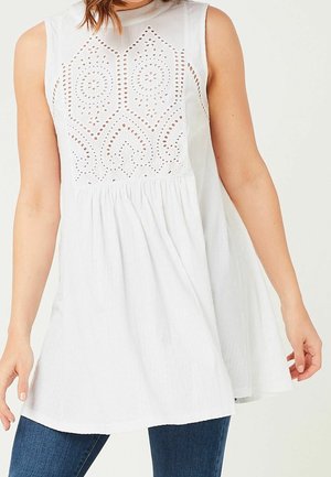 Ærmeløs hvid tunika-top med broderede hulmønstre på brystet, løs pasform, båret med mørkeblå jeans, vist fra midt på kroppen til hofterne.