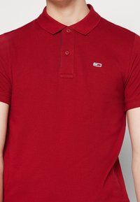 Červená polo košile s texturovanou tkaninou, zapínací límec a krátké rukávy. Na levé hrudi se nachází malé vyšívané logo.