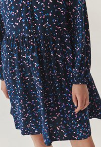 Bloemenjurk met een diepblauwe achtergrond en verspreide blauwe en roze bladmotieven. Lange mouwen met elastische manchetten. Knopdetails.