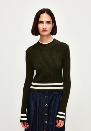 Rippstrickpullover in dunklem Olivgrün mit hohem Kragen und gestreiftem weißen Saum sowie Ärmelakzenten, kombiniert mit einem A-Linien-Denimrock mit goldenen Knöpfen.