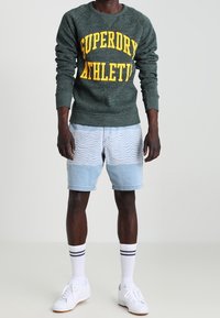 Sudadera verde moteada con "SUPERDRY ATHLETIC" en amarillo, combinada con pantalones cortos de mezclilla azul claro con un patrón ondulado. Zapatillas blancas y calcetines.