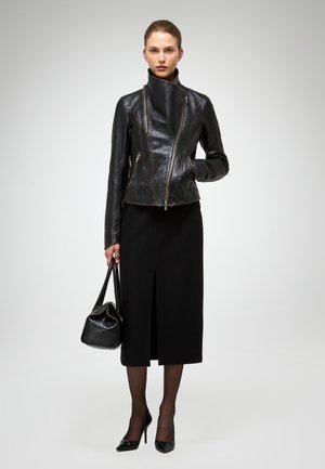 Sieviete valkā melnu asimetrisku ādas jaku, melnu midi svārku ar priekšējo šķēlumu, melnas caurspīdīgas zeķubikses, smailus papēžus un tur melnu somiņu.