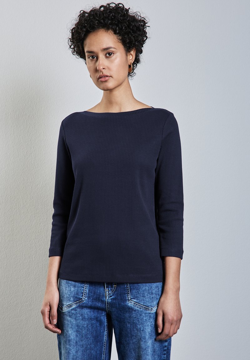 Marineblaues geripptes Langarmoberteil mit weitem Ausschnitt, kombiniert mit hellblauen Jeans. Der Stoff hat eine glatte Textur und eine figurbetonte Silhouette.