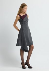 Kleid in Anthrazitgrau mit burgunderrotem dreieckigem Ausschnitt, asymmetrischem Saum, weicher Textur und schlankem Schnitt. Schwarze High-Heelsandalen.