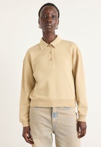Pullover beige con colletto, tre bottoni e polsini a costine. Presenta un logo in rilievo sul petto e un design dalla vestibilità rilassata.