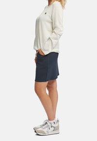 Chemise beige clair à manches longues avec un petit logo brodé, associée à une jupe bleu foncé et des baskets gris-blanc.