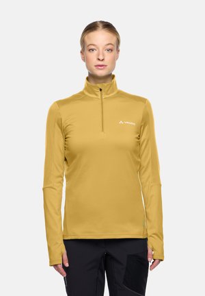 Woman wearing mustard yellow half-zip sports top with Vaude logo and black pants, standing against a plain light background.