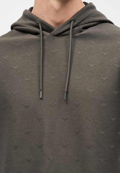 Κοντινό πλάνο ενός ατόμου που φοράει γκρι σκούρο φούτερ με κουκούλα, με διακριτικά επαναλαμβανόμενα ανάγλυφα λογότυπα πουλιών και κορδόνια να κρέμονται προς τα κάτω.