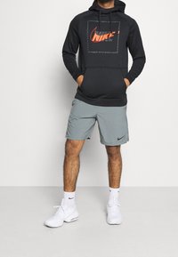 Felpa nera Nike con logo rosso, abbinata a pantaloni corti grigi. Sneakers bianche e calzini alla caviglia completano l'outfit, mostrando uno stile sportivo.