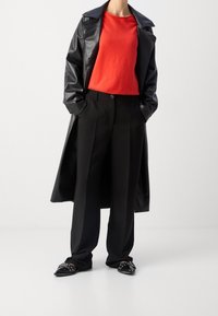 Long manteau en cuir noir, pull rouge vif, pantalon noir et chaussures noires à bout pointu avec détails décoratifs en métal.