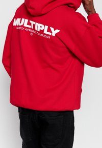 Sudadera roja con capucha que presenta un gran logo blanco de "MULTIPLY" en la parte posterior, hecha de una tela suave, con un corte relajado y un bolsillo tipo canguro.