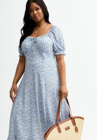 Robe bleu clair avec un motif floral, manches bouffantes et encolure à cordon ; associée à un sac beige avec des accents marron.