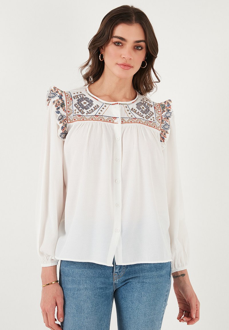 Witte blouse met borduursel langs de schouderpas en gerimpelde schouders. Heeft een knoopsluiting en lange mouwen, gecombineerd met blauwe jeans.