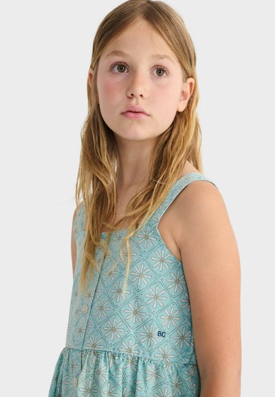 Mladé dievča s dlhými blond vlasmi má na sebe modré šaty bez rukávov s bielymi vzormi margarét, pozerá sa mierne nahor a do strán na jednoduchom pozadí.