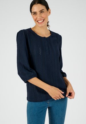 Blusa blu navy con un motivo testurizzato, con maniche a tre quarti, collo rotondo e dettagli con bottoni frontali. Indossata sopra dei jeans di denim.