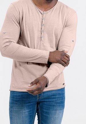 Tmavě pigmentovaný člověk nosící béžové henley tričko s dlouhým rukávem a modré džíny, upravující levý rukáv pravou rukou.