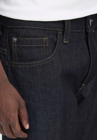 Jeans in denim scuro con cuciture gialle a contrasto, design a cinque tasche e hardware argentato. Primo piano delle tasche laterali e frontali.