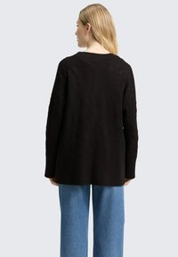 Zwarte gebreide trui met een losse pasvorm, geribbelde manchetten en zoom. Draagt lichtblauwe wijd uitlopende jeans. Gezien vanaf de achterkant tegen een effen achtergrond.