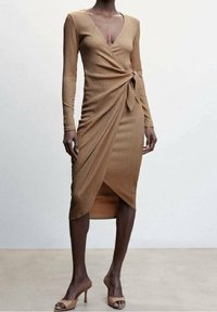 Femme portant une robe midi enveloppante à manches longues de couleur camel, associée à des chaussures à talons ouverts de la même teinte, se tenant devant un fond uni.