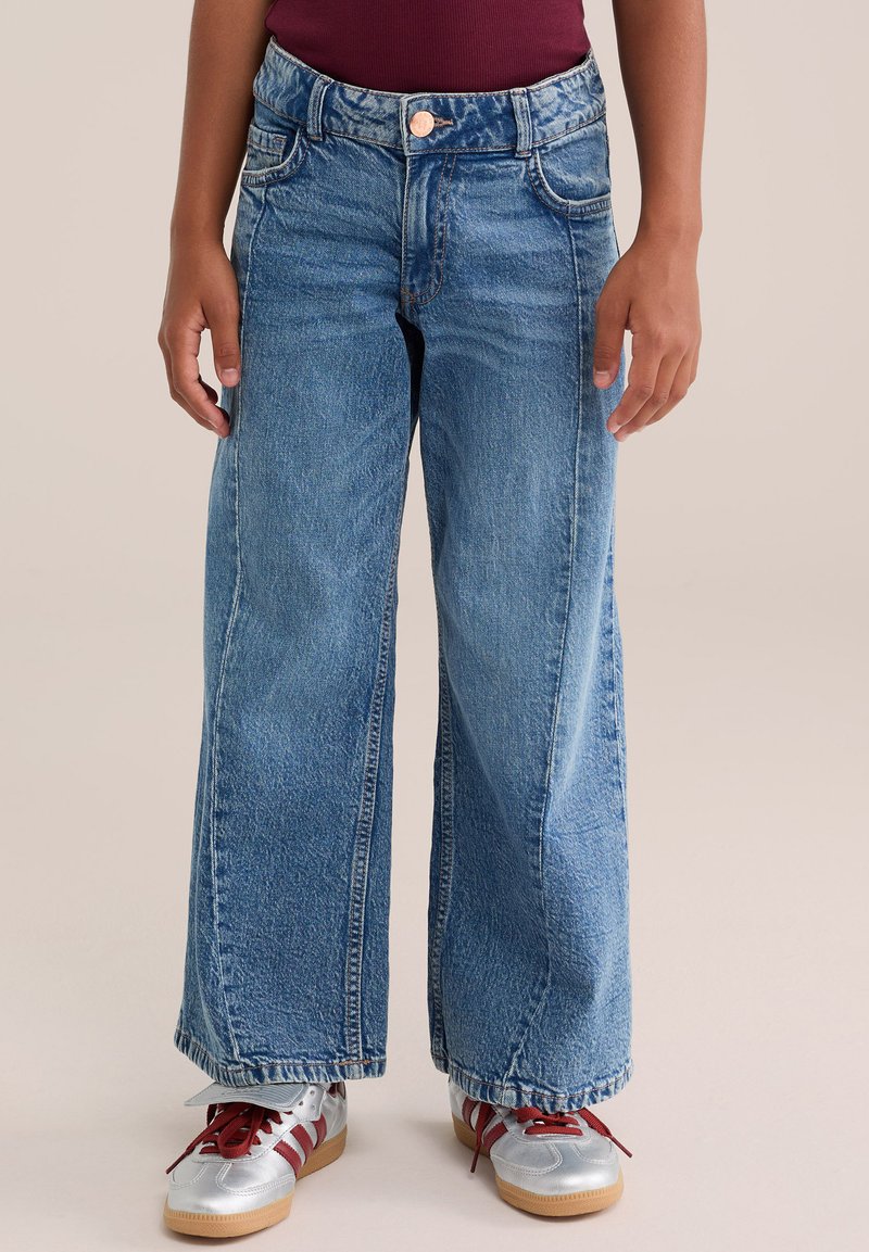 Enfant portant un jean bleu à jambes larges avec poches avant, un haut bordeaux et des baskets argentées avec des lacets rouges et des rayures sur un fond neutre.