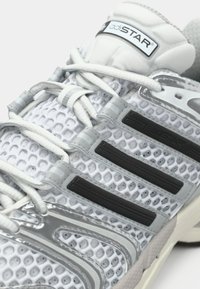 Bijela sportska cipela s mrežastim gornjim dijelom, sivim akcentima, crnim teksturiranim prugama i reflektirajućim detaljima. Ima podstavljeni potplat i sustav vezivanja.