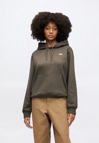 Olivovo zelená mikina s kapucňou so sťahovacou šnúrkou, predným vreckom a bielym logom na hrudi. Nosená s béžovými nohavicami, má voľný strih a mäkkú textúru.