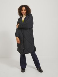 Manteau matelassé noir avec capuche, présentant une fermeture éclair à l'avant, des poches latérales et des manches longues, porté sur un pull en tricot et un jean sombre.
