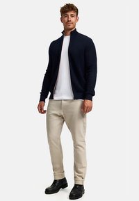 Tengerészkék zipzáras pulóver texturált mintával, fehér póló fölött viselve, bézs nadrággal és fekete cipővel párosítva. Laza szabás, minimalista design.