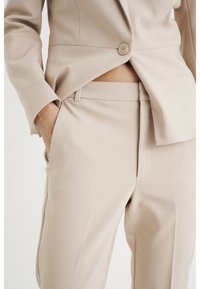 Traje entallado beige con blazer ajustado y pantalones de pierna recta. Tela suave, cierre de un botón, bolsillos laterales en los pantalones. Textura lisa.