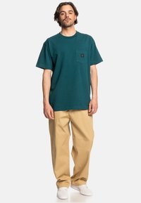Tyrkysové bavlněné tričko s kapsou na hrudi. Spárováno s béžovými kalhotami volného střihu a bílými teniskami, což vytváří neformální outfit.