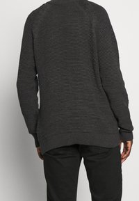 Person, der einen dunkelgrauen strukturierten Strickpullover und schwarze Hosen trägt, von hinten vor einem schlichten hellen Hintergrund gezeigt.