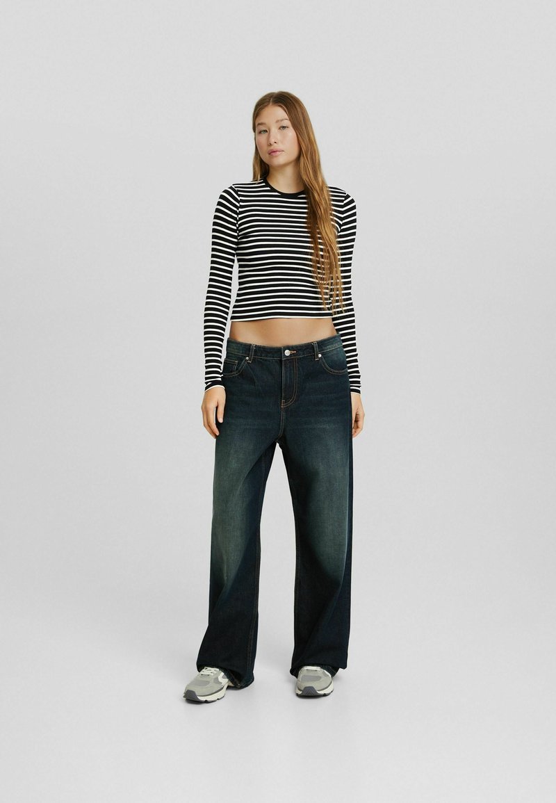 Schwarz-weißer gestreifter Langarm-Crop-Top kombiniert mit dunkelblauen weiten Jeans und grauen Sneakers, stehend vor einem schlichten Hintergrund.